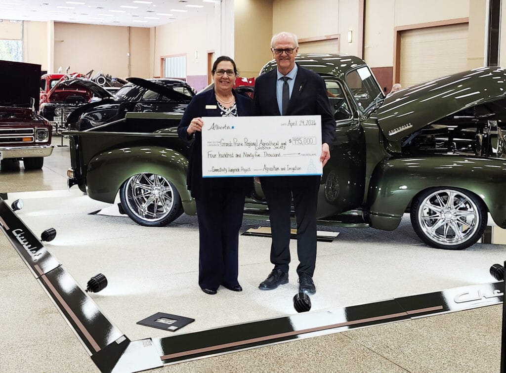 A man and a woman smile while holding a large check for four hundred ninety five thousand dollars, surrounded by several polished classic cars, including a dark green pickup truck, at an indoor event.