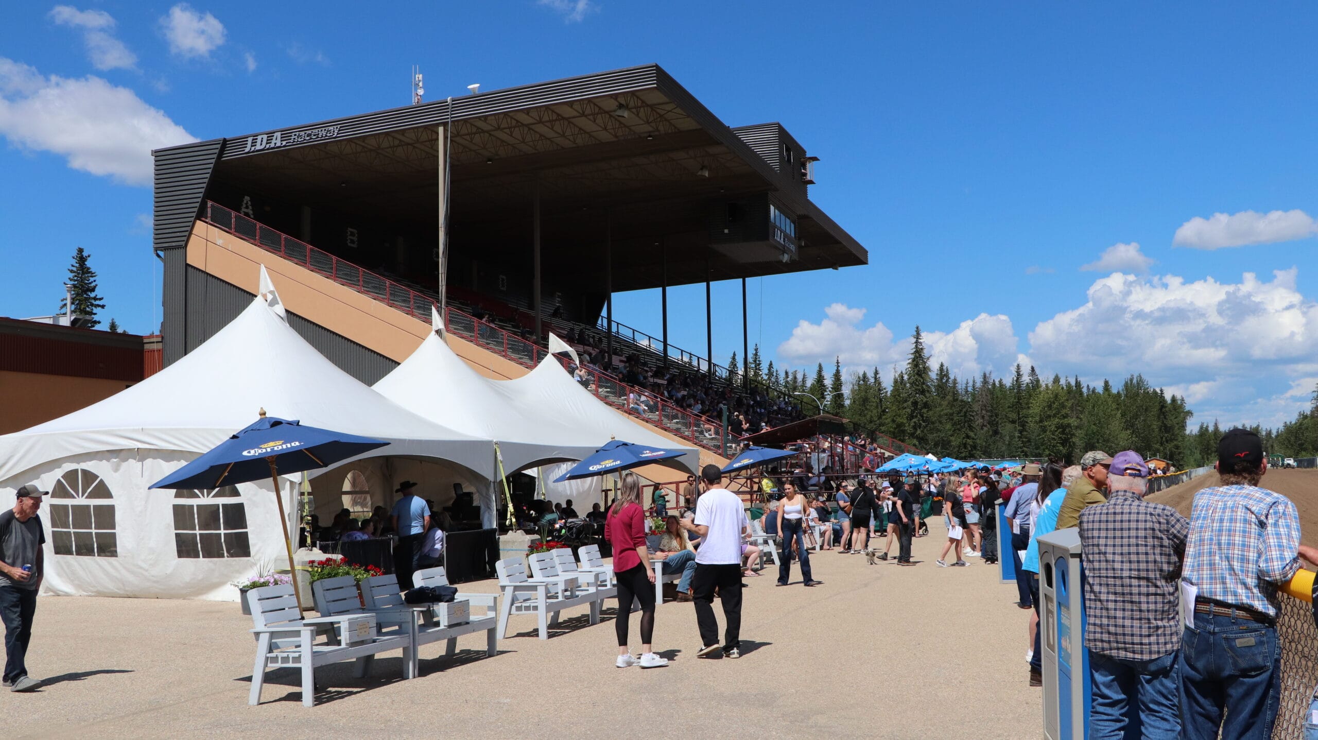 Many people are gathered near white tents and blue umbrellas on a sunny day at JDA Raceway, with spectators visible in the large grandstand overlooking a dirt track.