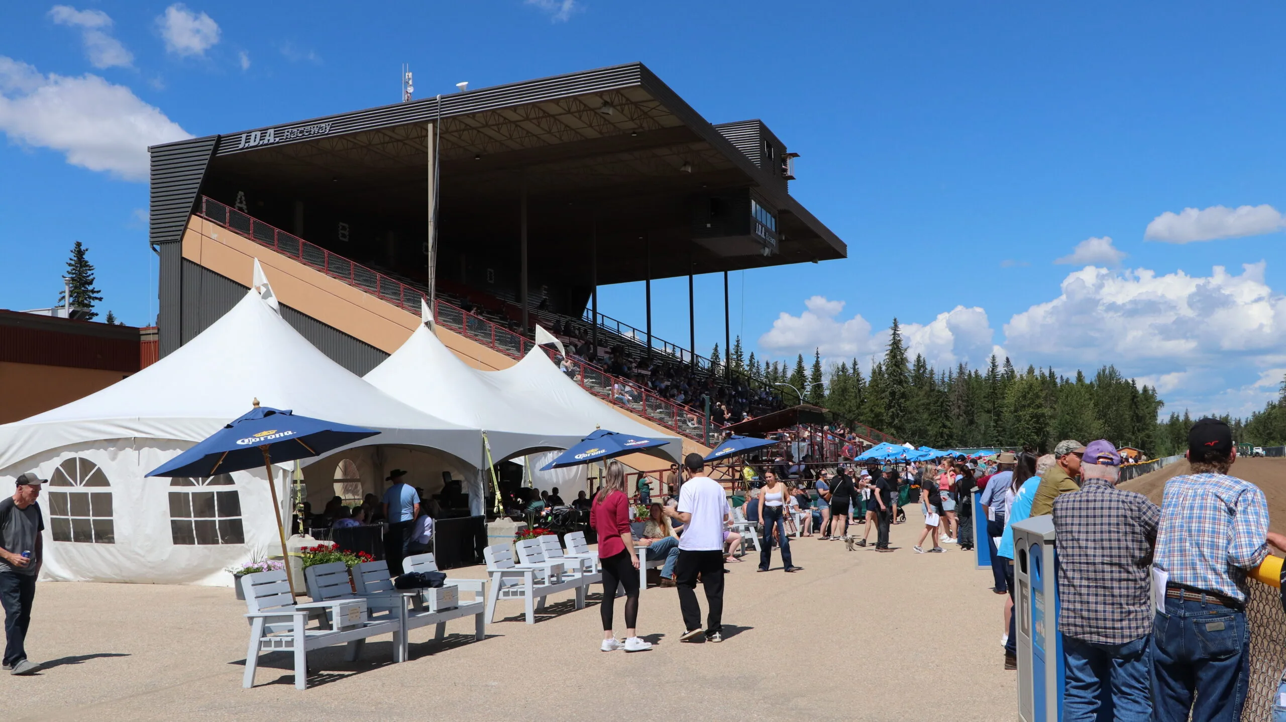 Numerous people are gathered around white tents and under a large grandstand at an outdoor raceway, all beneath a bright blue sky.