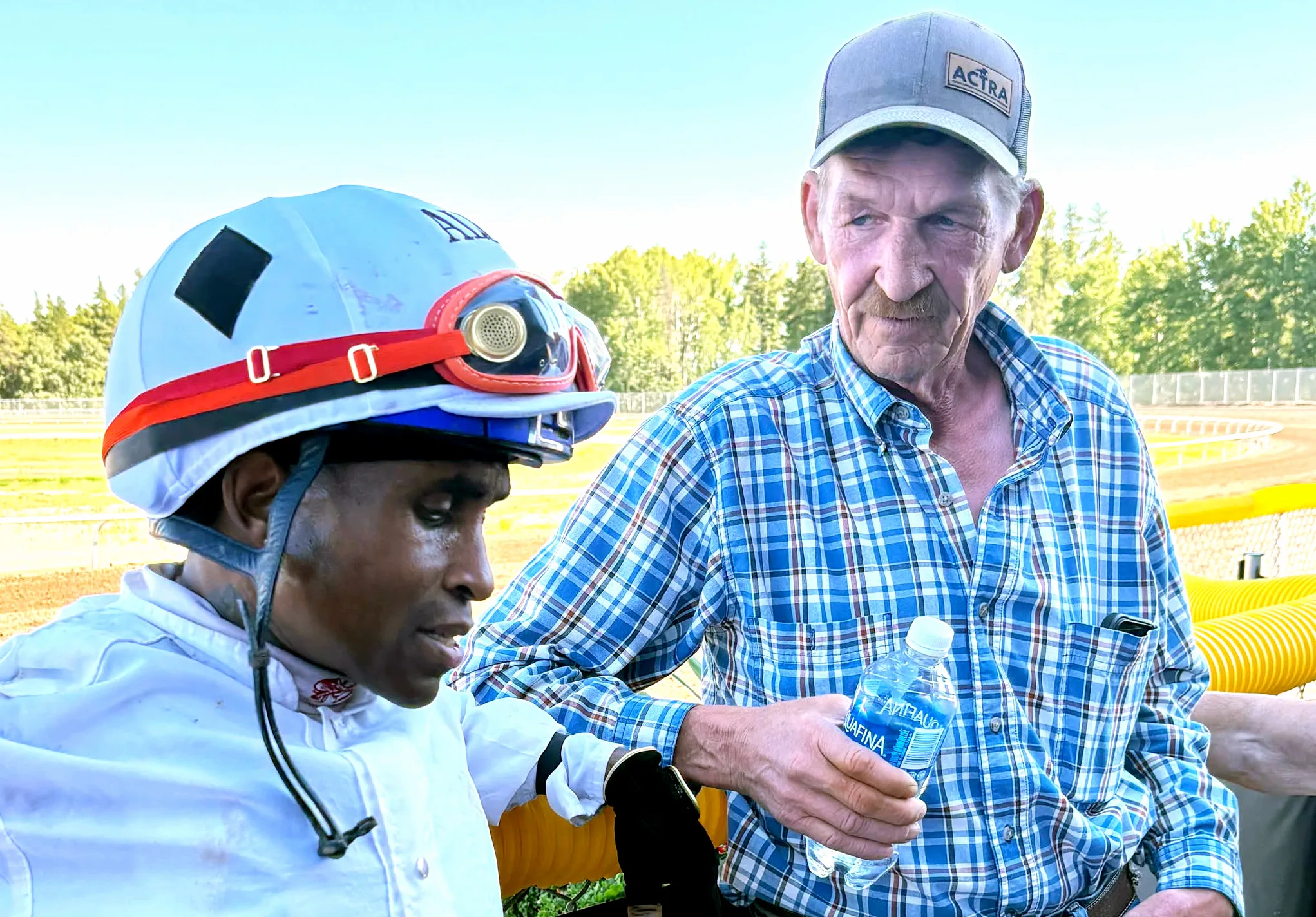 An older man in a plaid shirt and baseball cap holds a bottle of water while looking at a jockey wearing a white uniform and helmet, both standing outdoors near a track.