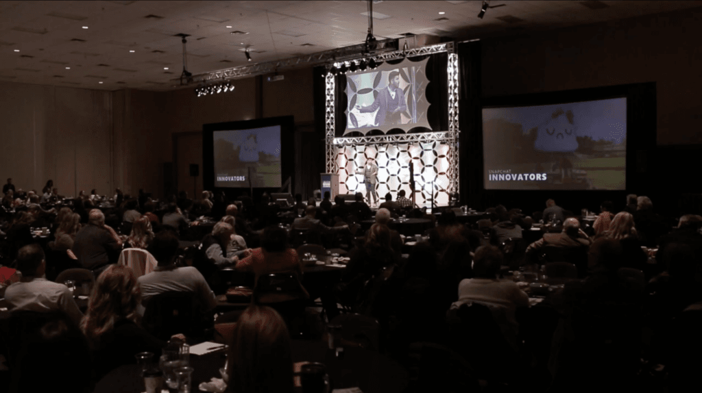 An audience sits at tables in a large, dimly lit conference room, facing a stage where a speaker presents before a central screen showing a man, flanked by two large projection screens displaying Snapchat Innovators.