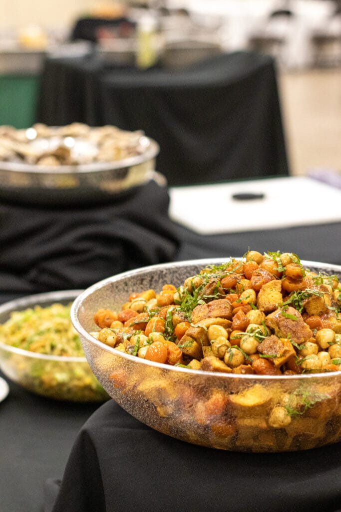 A large glass bowl overflowing with roasted tomatoes, chickpeas, and bread chunks topped with green herbs sits on a black tablecloth, with other serving dishes visible in the blurry background.