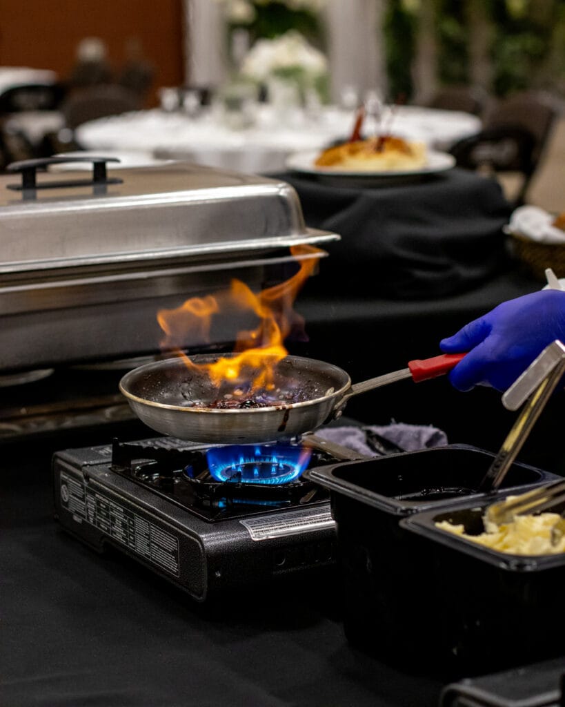 A chef in a blue glove cooks food in a flaming pan on a gas burner at an event, with blurry dining tables in the background.
