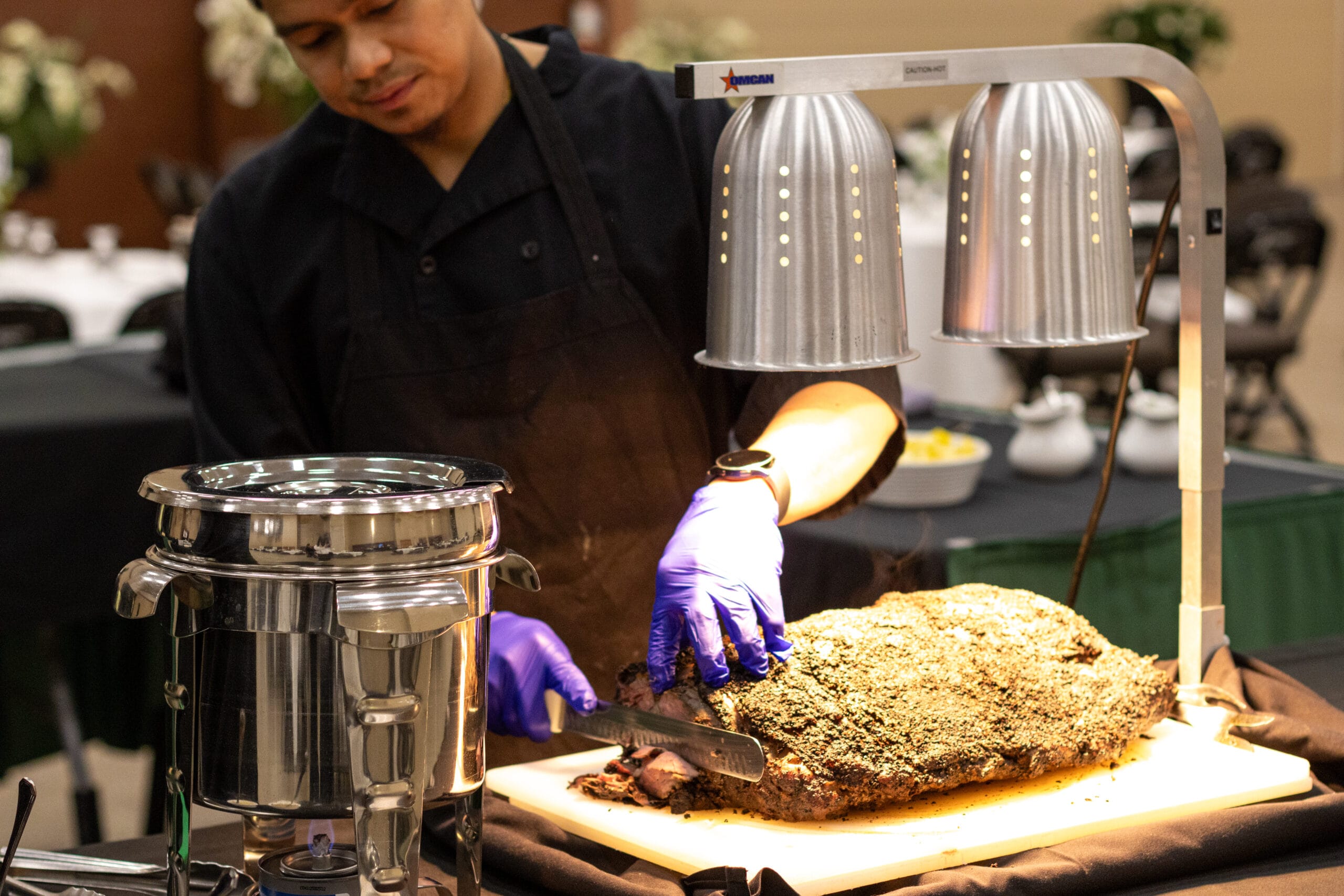 A chef wearing blue gloves carves a large seasoned roast on a buffet table beneath heat lamps.