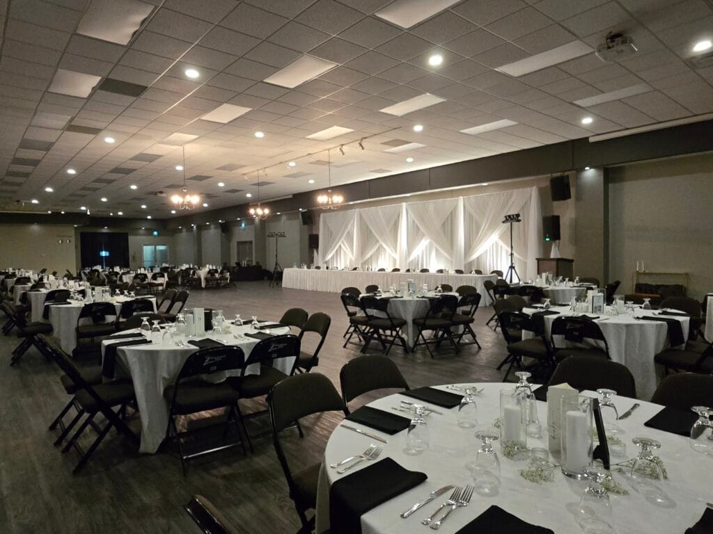 A large, well lit event hall is prepared with numerous round and rectangular tables featuring white tablecloths, dark chairs, and elegant place settings adorned with candles and small flowers.
