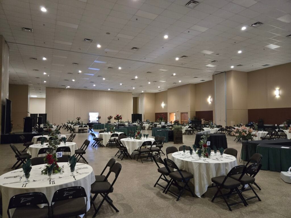 A spacious event hall is meticulously arranged with numerous round and rectangular tables draped in various colored cloths, set with chairs, place settings, and floral centerpieces, illuminated by bright overhead and wall lighting.