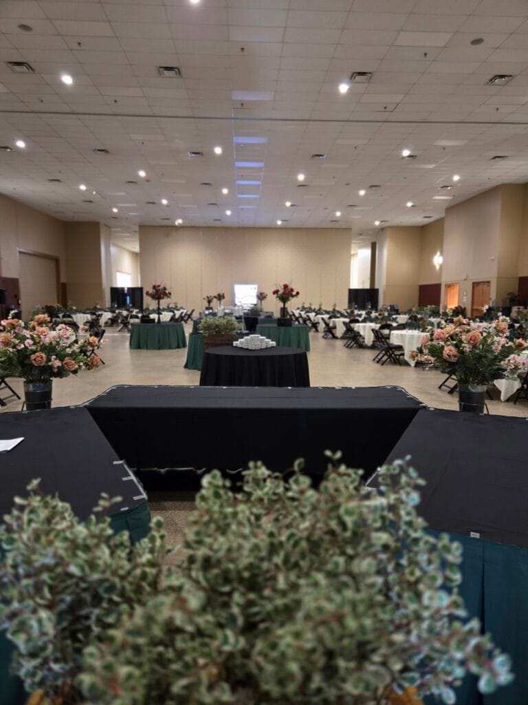 An expansive event hall is set with numerous tables, many adorned with floral arrangements and draped in various colored cloths, with green plants blurring in the foreground.