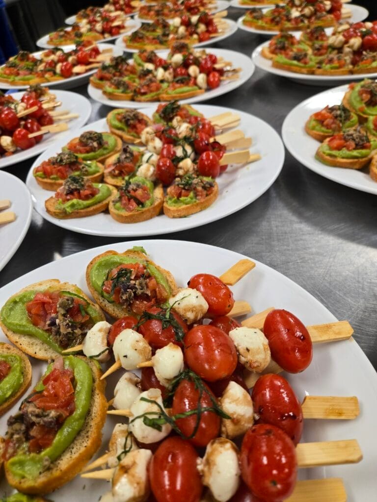 Numerous white plates are filled with avocado toast appetizers topped with tomato relish and basil, alongside skewers of cherry tomatoes and mozzarella balls, all arranged on a dark surface.